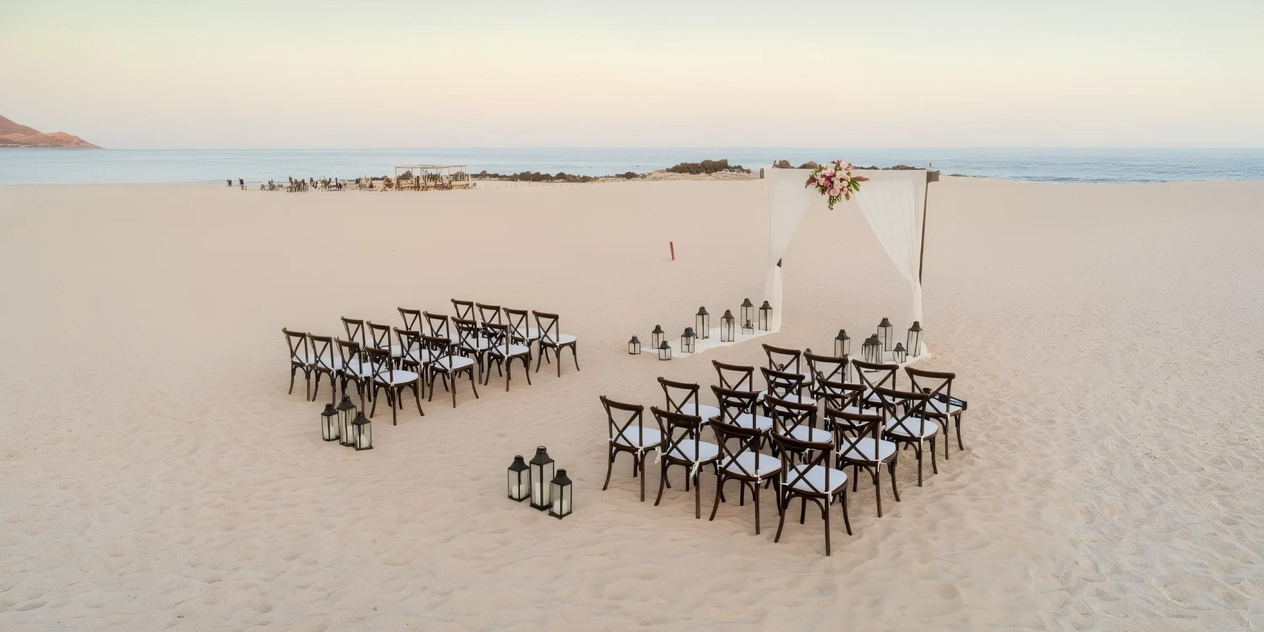 Beach wedding setup with wooden chairs and lanterns facing a draped arch adorned with flowers, near the ocean under a clear sky.