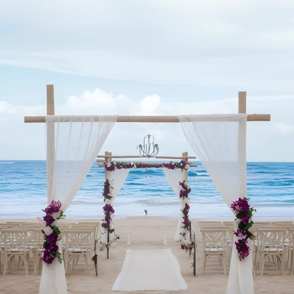 Wooden arch with white drapes and purple flowers decorates a beachside wedding setup, facing the ocean. Rows of white chairs line the sandy aisle.