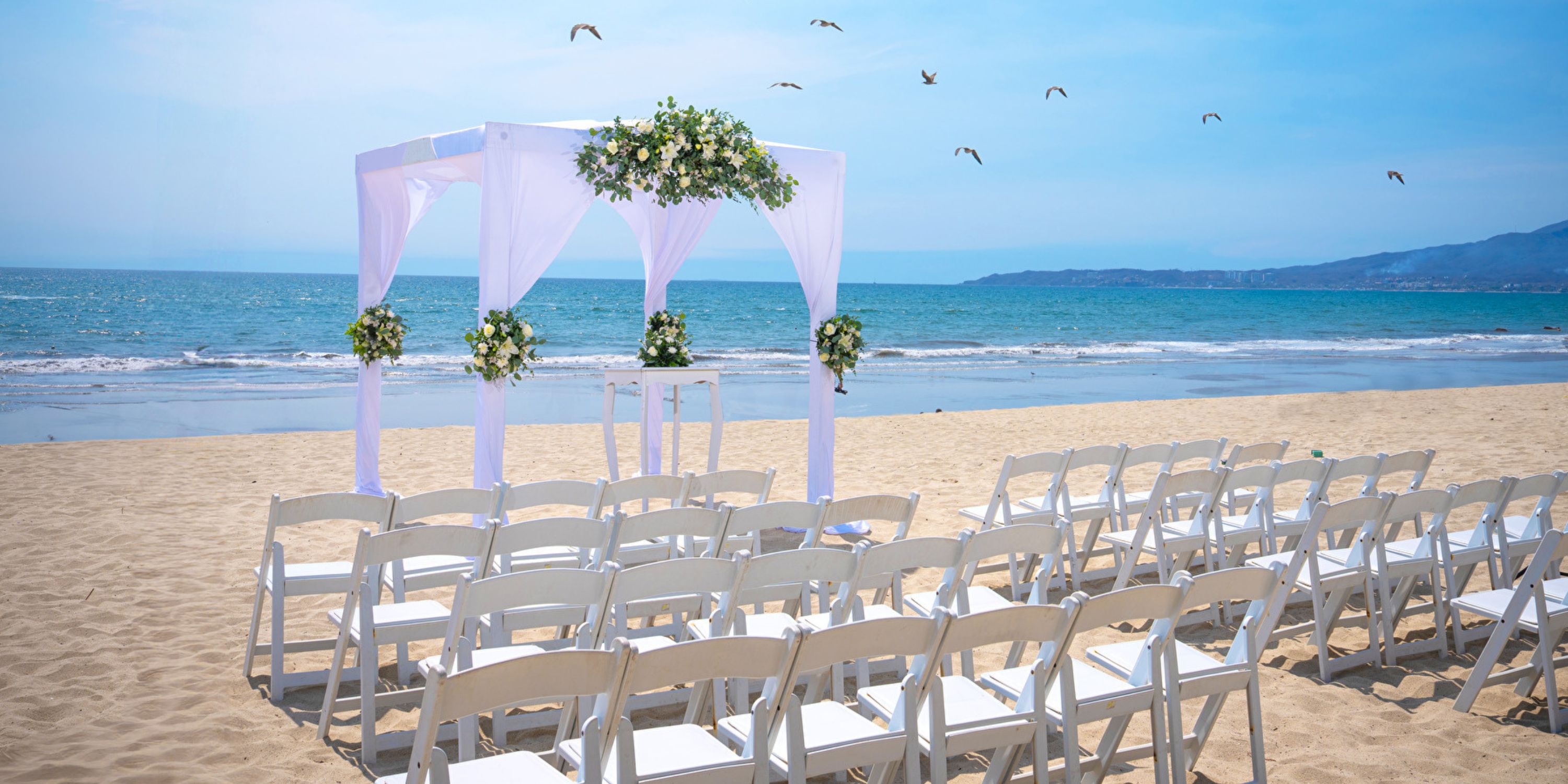 White chairs and a floral arch set up for a destination wedding on the beach at Hotel Riu Jalisco.