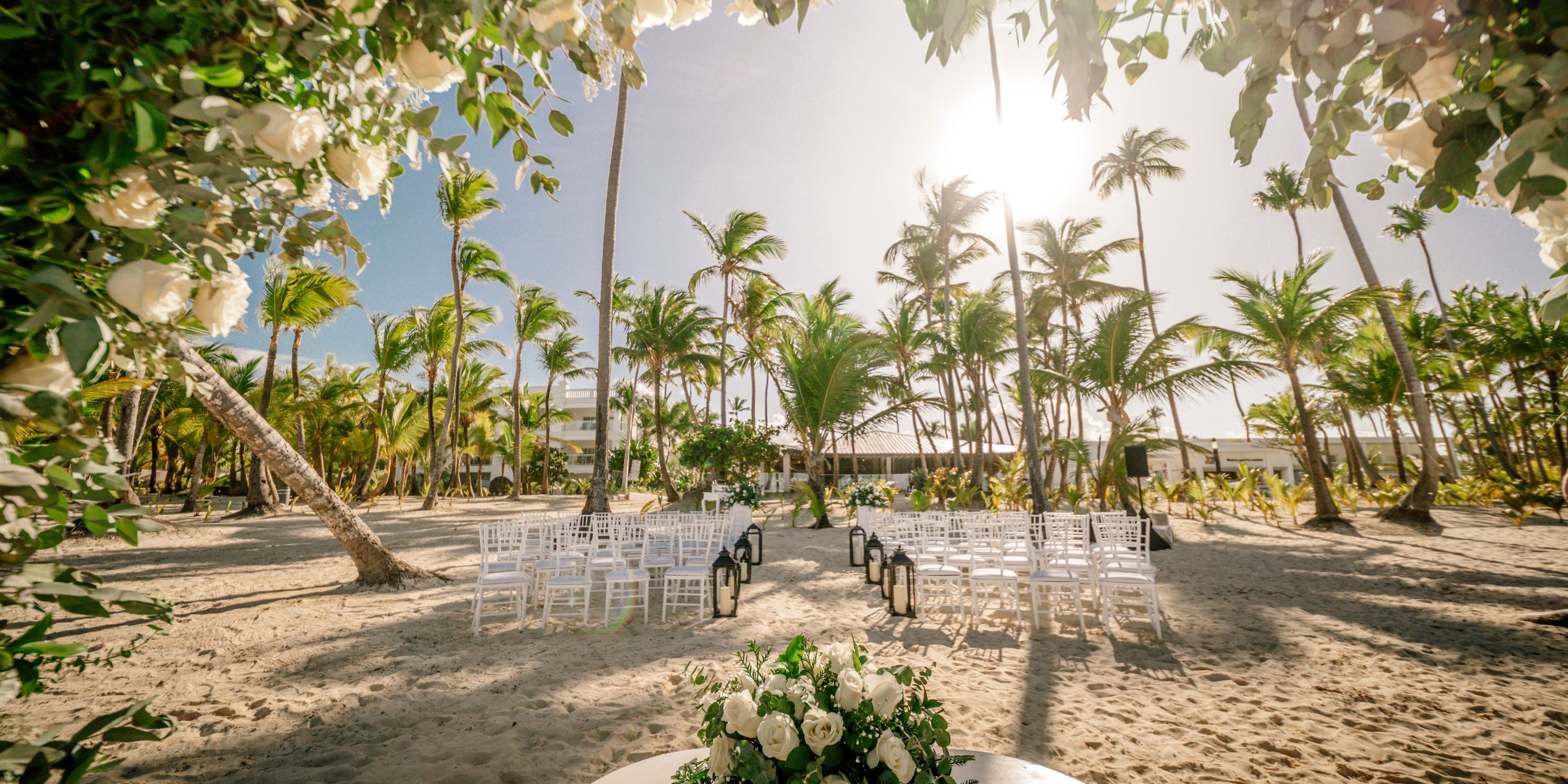 White chairs arranged for a destination wedding at Hotel Riu Bambu, shaded by palm trees on the beach.