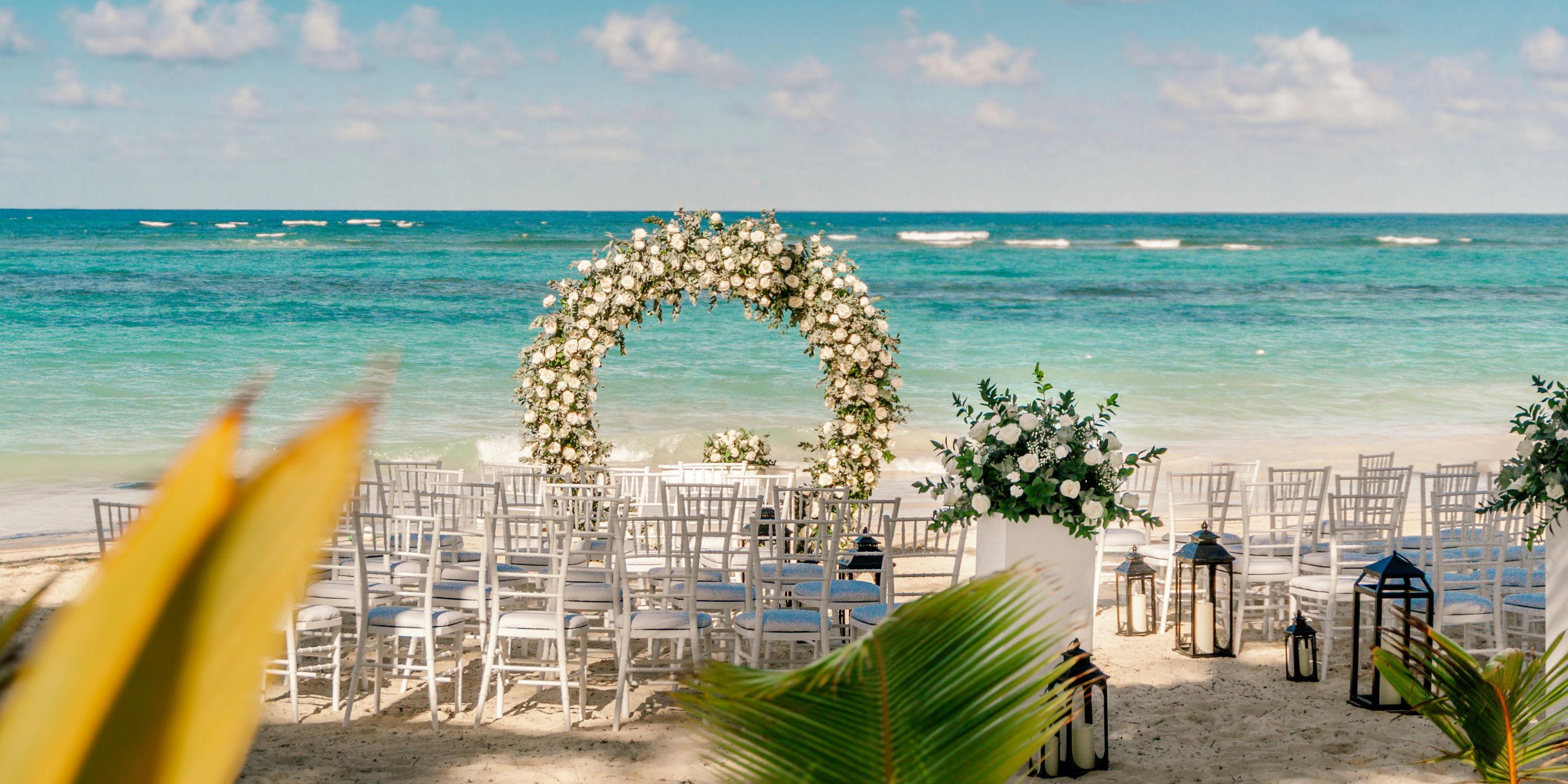 White chairs and a floral arch set up for a destination wedding on the sandy beach at Hotel Riu Bambu.