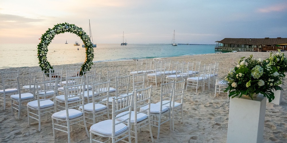 Rows of white chairs arranged for a destination wedding face a floral arch on the beach at Hotel Riu Palace Aruba.