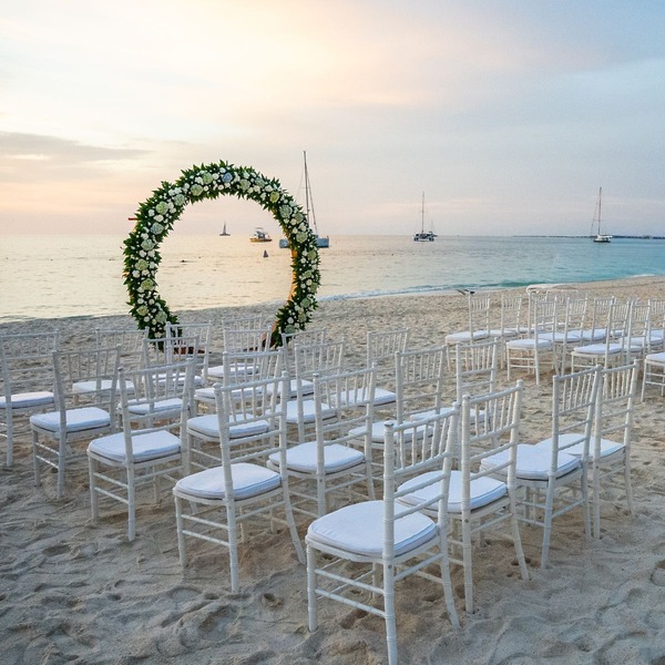 White chairs and a floral arch set up for a destination wedding at sunset, with boats behind.