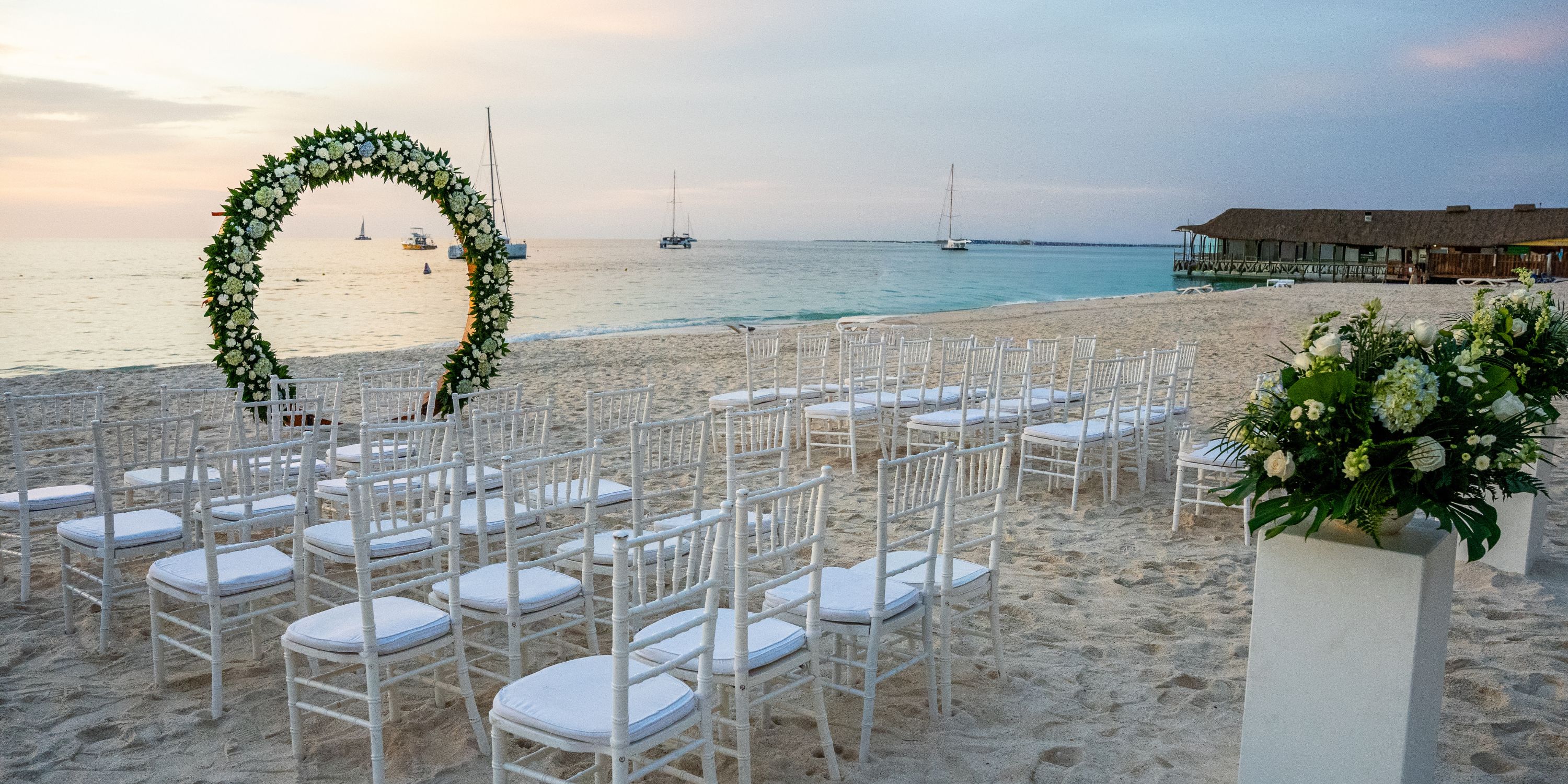 Rows of white chairs arranged for a destination wedding face a floral arch on the beach at Hotel Riu Palace Aruba.