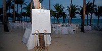 A wedding seating chart is displayed on the beach at Hotel Riu Palace Riviera Maya, set against a backdrop of palm trees—a perfect touch for a destination wedding.