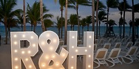 A large illuminated "R & H" glows at dusk on the beach of Hotel Riu Palace Riviera Maya, surrounded by palm trees—creating a picturesque backdrop for a destination wedding celebration.