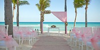 A stunning destination wedding at Hotel Riu Palace Riviera Maya, featuring elegant pink decor, white chairs, and a beautiful floral arch on the beach.
