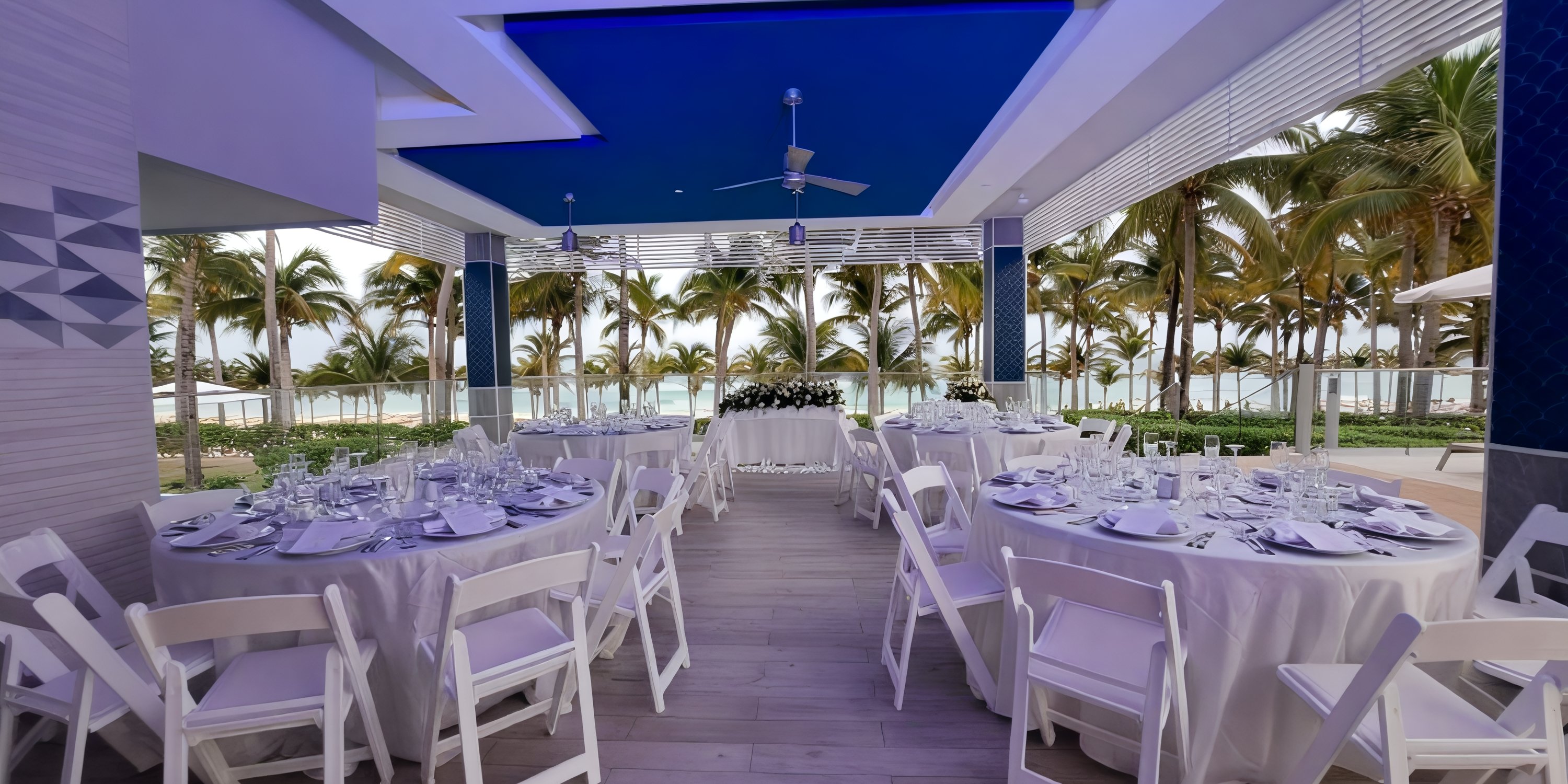 White tables arranged for a destination wedding event outdoors at Hotel Riu Palace Riviera Maya, near palm trees.