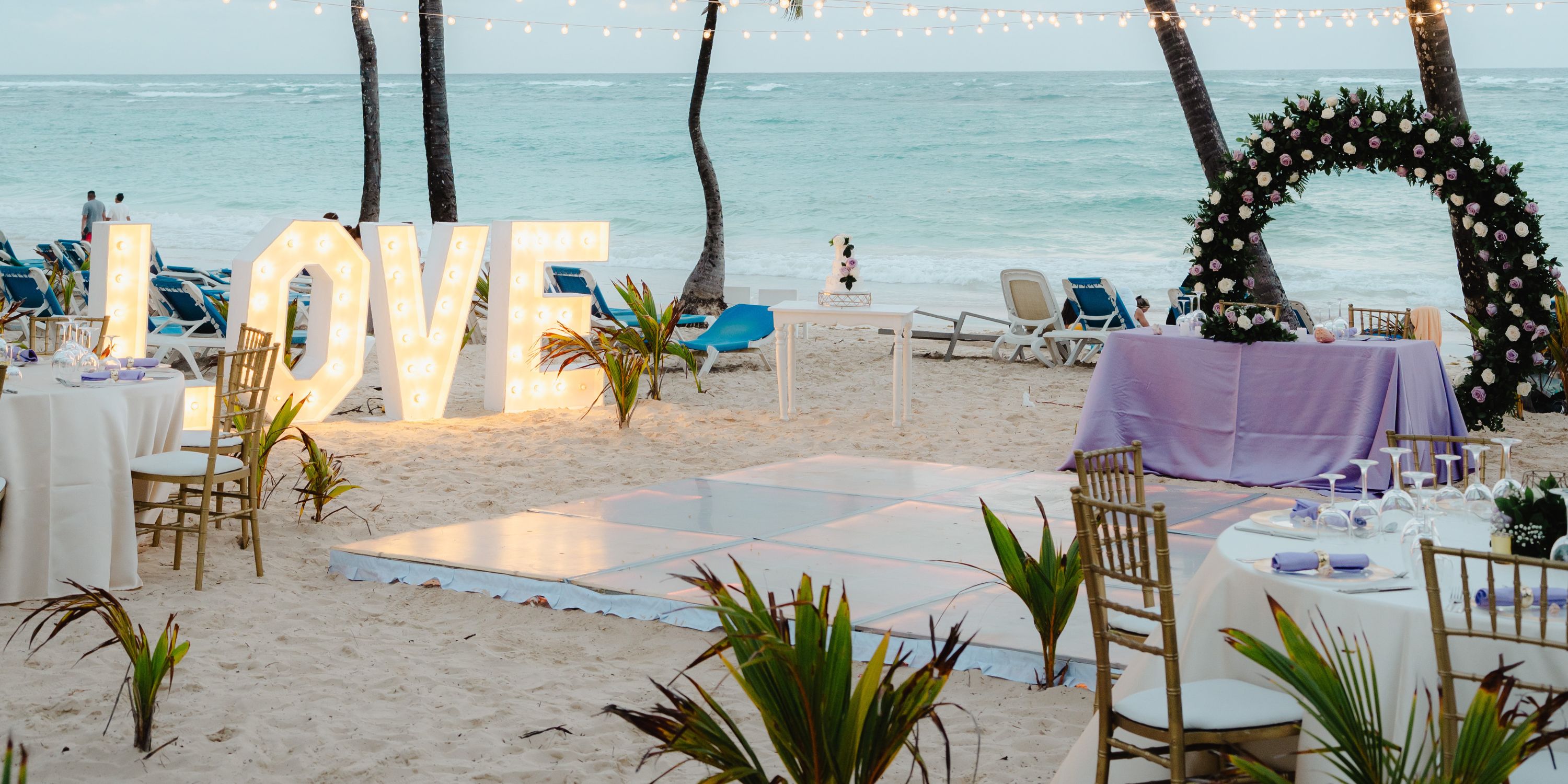 Destination wedding at Hotel Riu Bambu featuring a "LOVE" marquee, twinkling lights, and floral arch by the ocean.