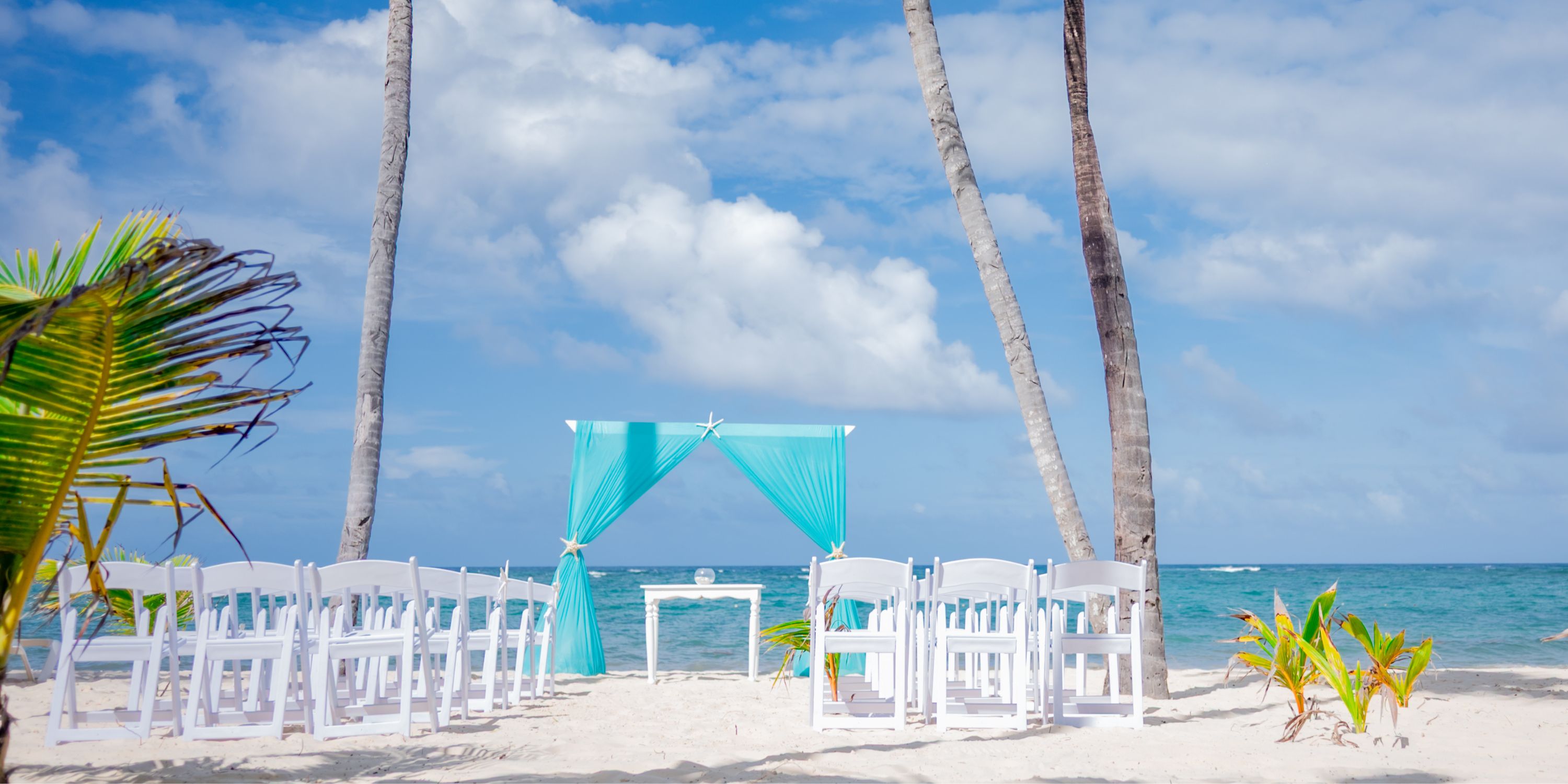 White chairs are set up for a destination wedding by a turquoise arch on the sandy beach at Hotel Riu Bambu.