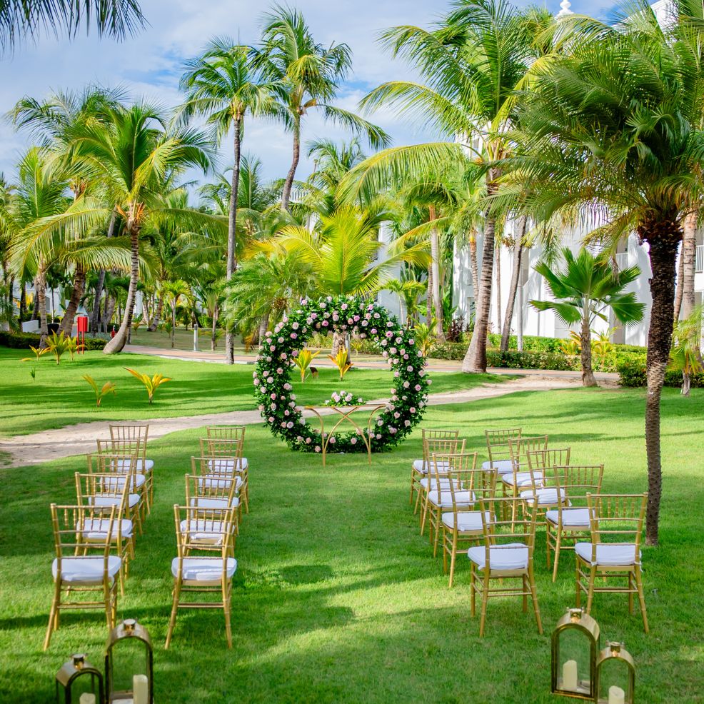 Gold chairs are arranged in rows at a destination wedding, facing a circular floral arch outdoors.