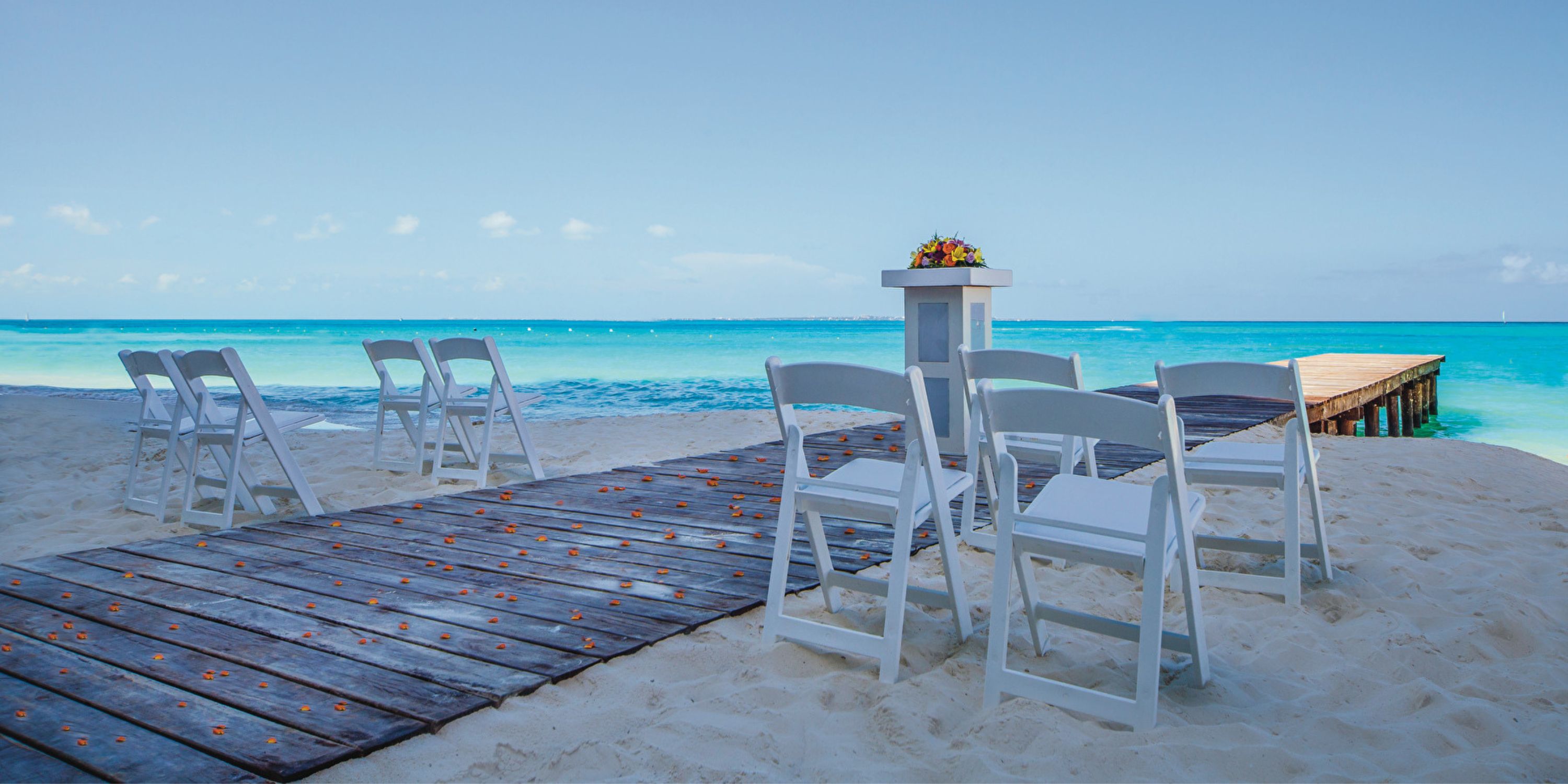 White chairs set for a destination wedding at Hotel Riu Cancun, with a wooden walkway and pier nearby.