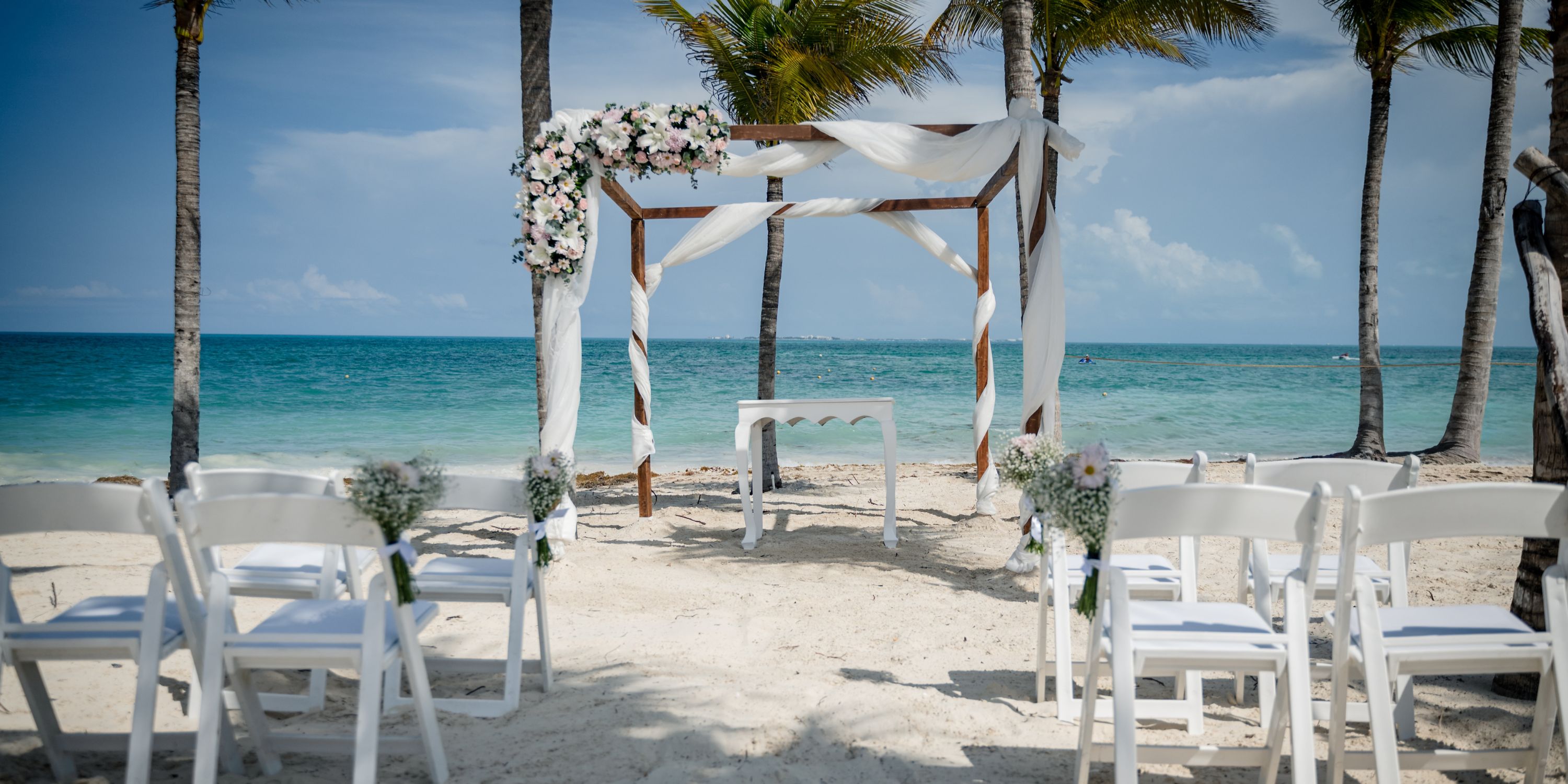 A destination wedding altar with white chairs set up on the sandy beach at Hotel Riu Dunamar by the ocean.