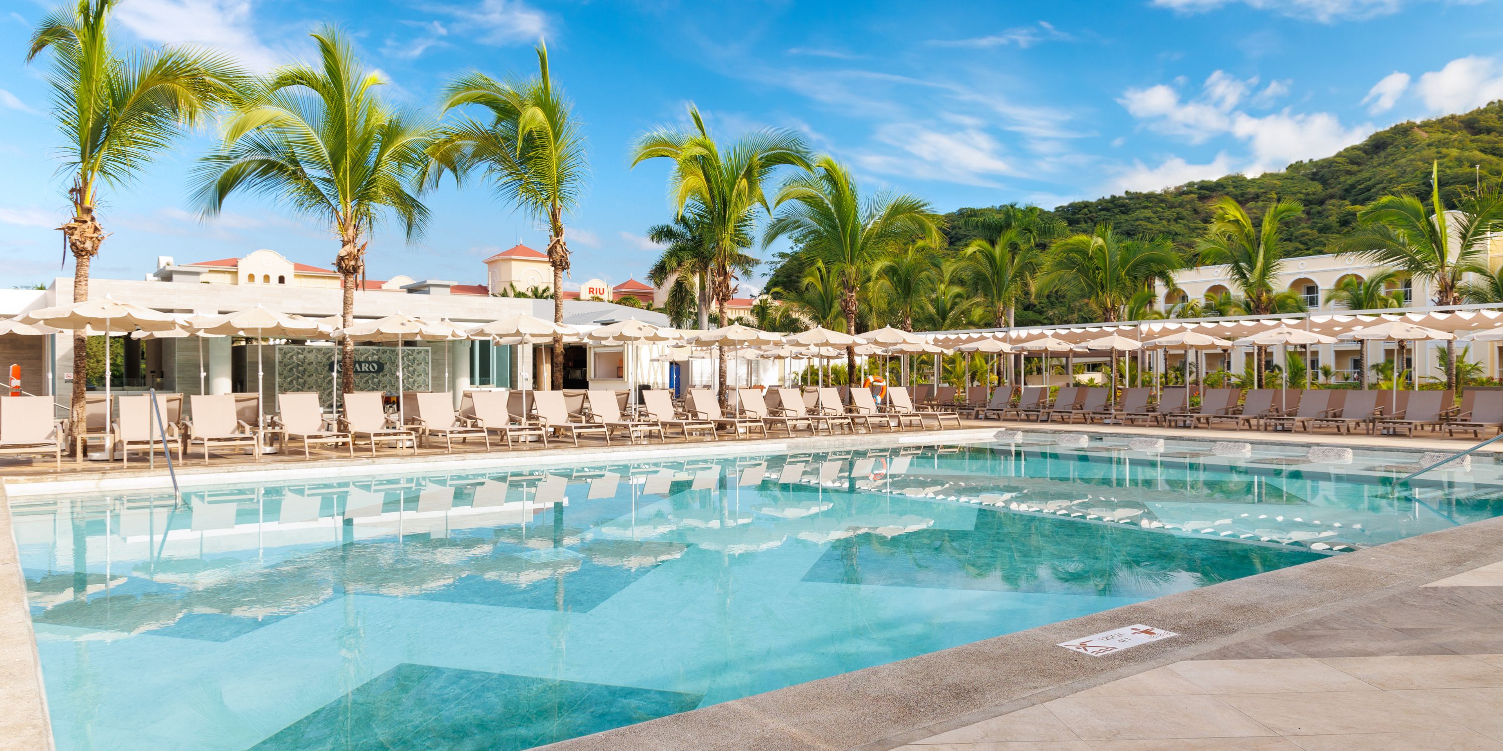 Hotel Riu Guanacaste offers a stunning setting for a destination wedding, featuring an inviting outdoor pool surrounded by lounge chairs and palm trees beneath a clear blue sky.