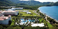 Aerial view of Hotel Riu Guanacaste, a coastal resort featuring several large buildings, a pool area with a winding design, and lush green hills in the background, adjacent to a curved beach shoreline.