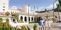 Four people stroll past the Hotel Riu Guanacaste, surrounded by lush tropical plants and palm trees in the foreground.