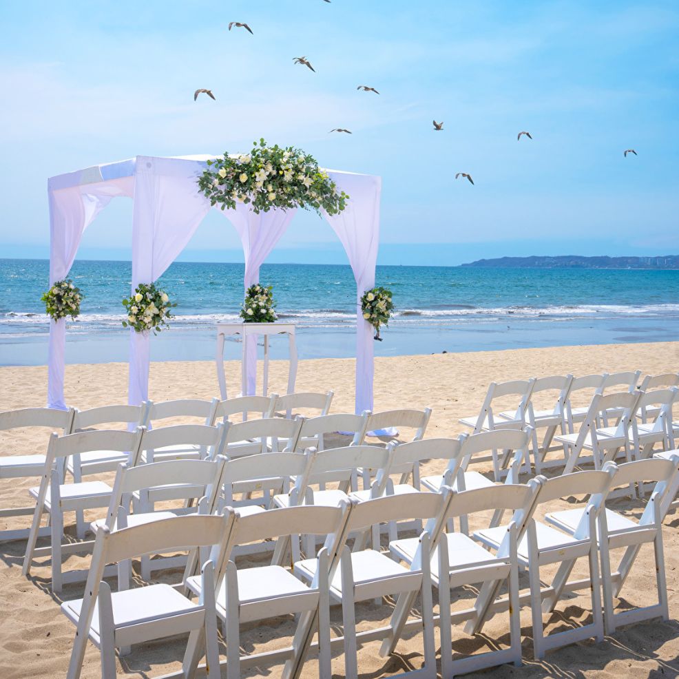 White chairs lined up on sandy beach face a decorated arch, ready for a destination wedding by the ocean.