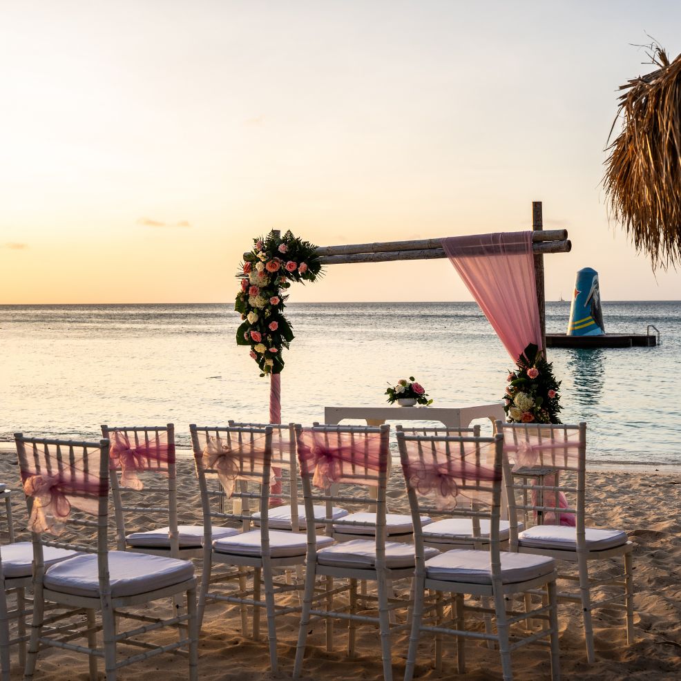 White chairs and a floral arch await guests at a sunset destination wedding set up on the beach.