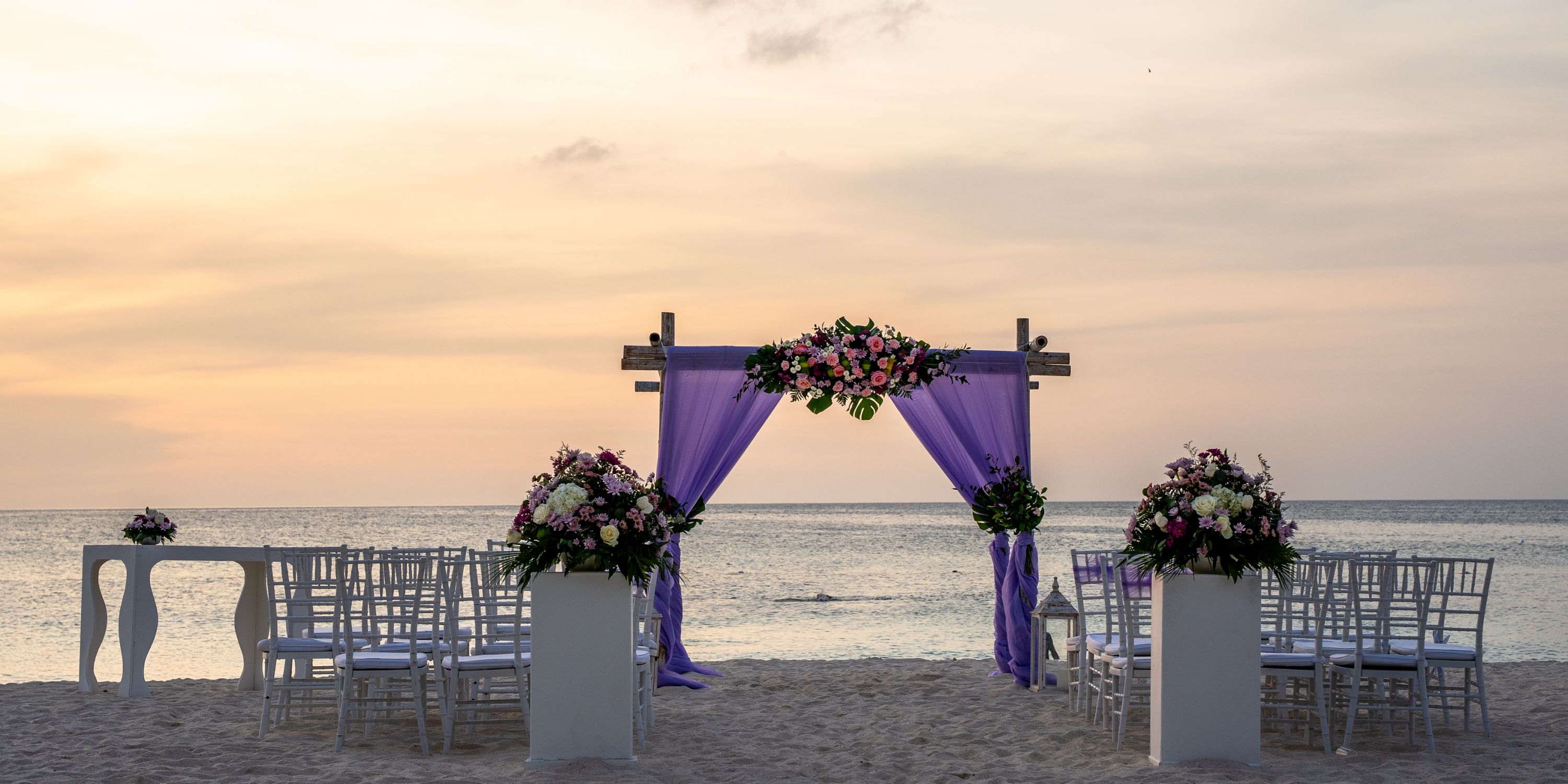 Destination wedding setup on the beach at Hotel Riu Palace Aruba with white chairs and a purple floral arch.