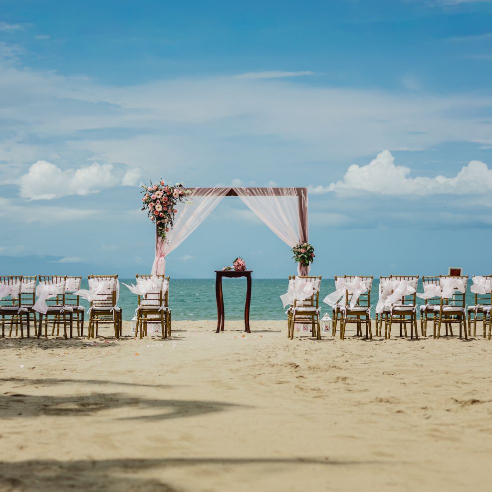A destination wedding setup on sandy beach with chairs, floral arch, and decorations facing the ocean.