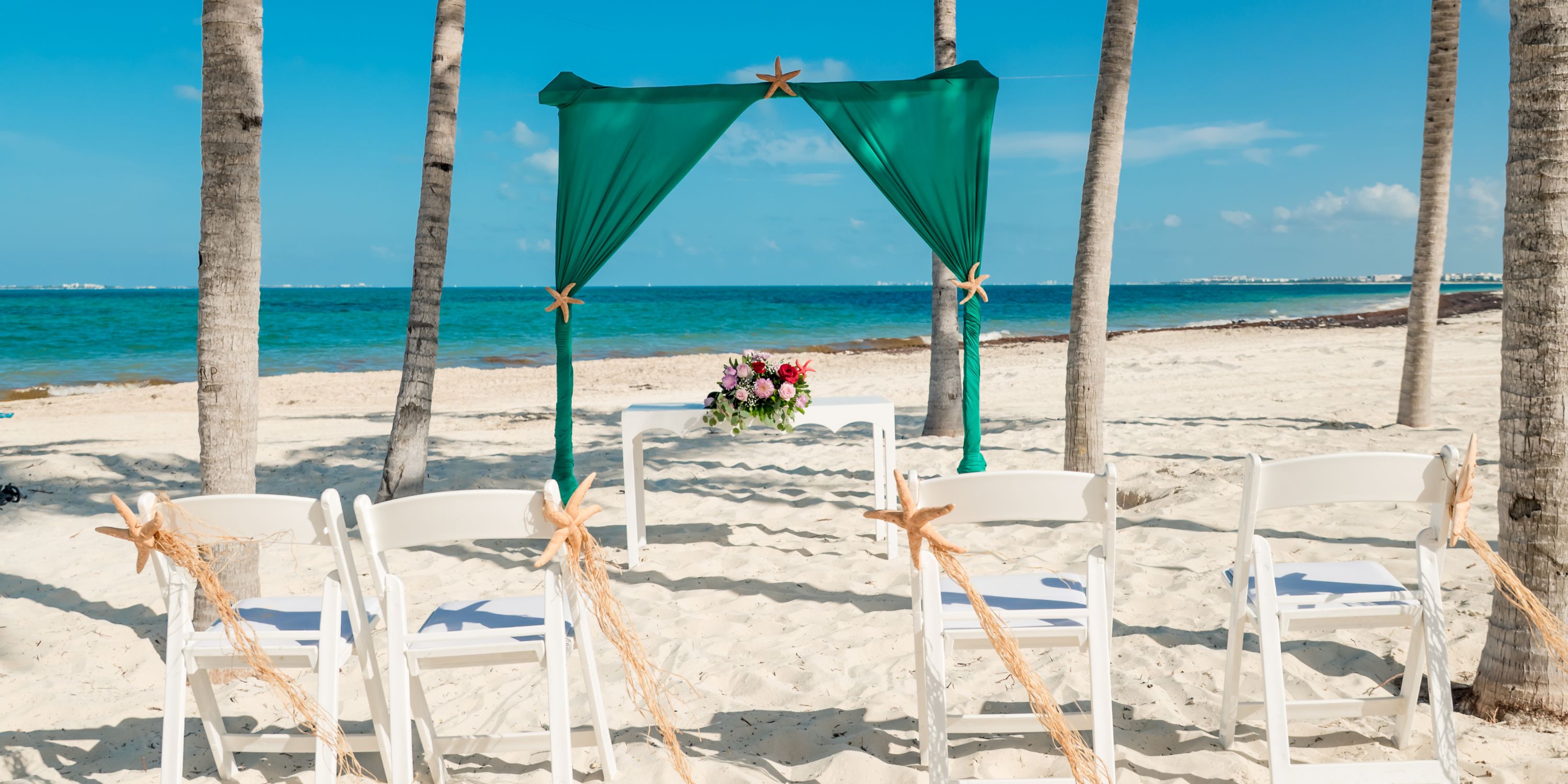 White chairs and a green arch arranged for a destination wedding at Hotel Riu Palace Costa Mujeres beach.