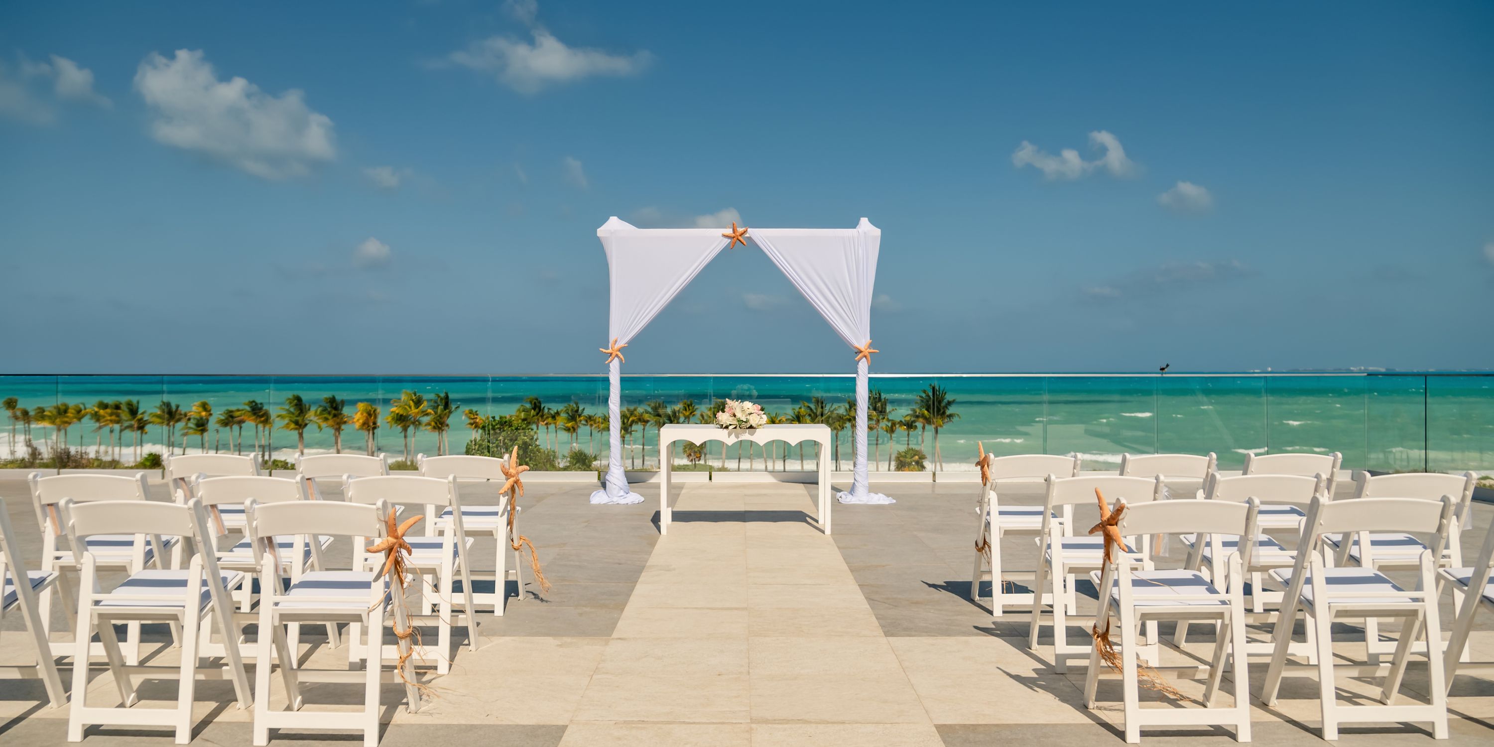 White chairs and a wedding arch set up for a destination wedding on the oceanfront terrace at Hotel Riu Palace Costa Mujeres.