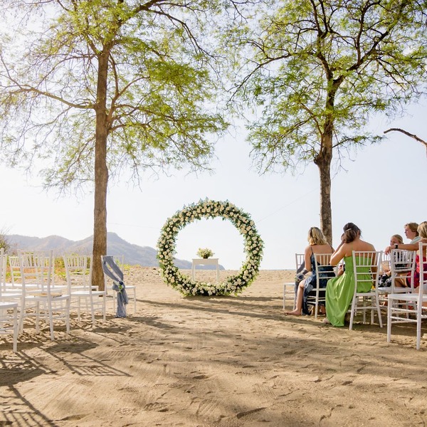 Destination wedding setup on the beach with white chairs facing a circular floral arch between two trees.