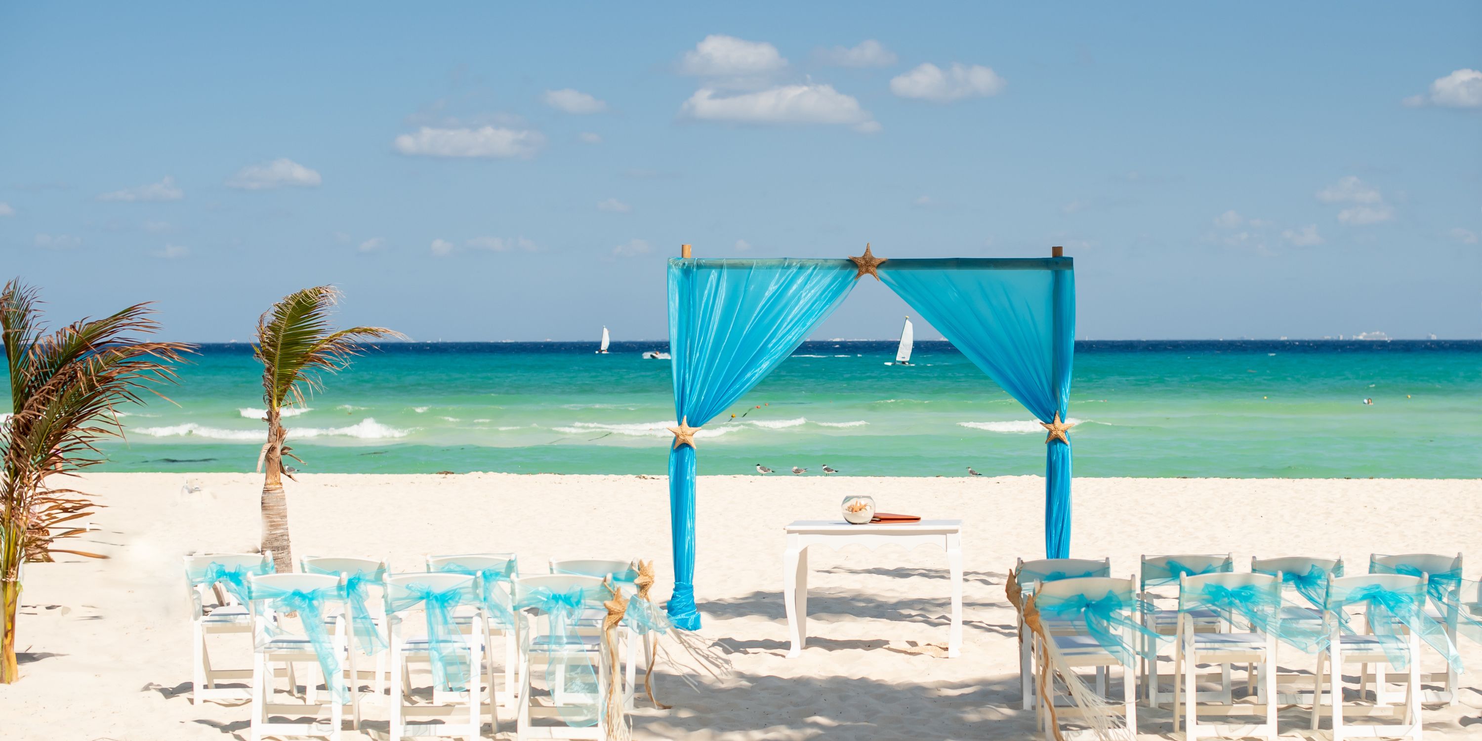White chairs and a blue arch arranged on the beach for a destination wedding at Hotel Riu Palace Mexico.