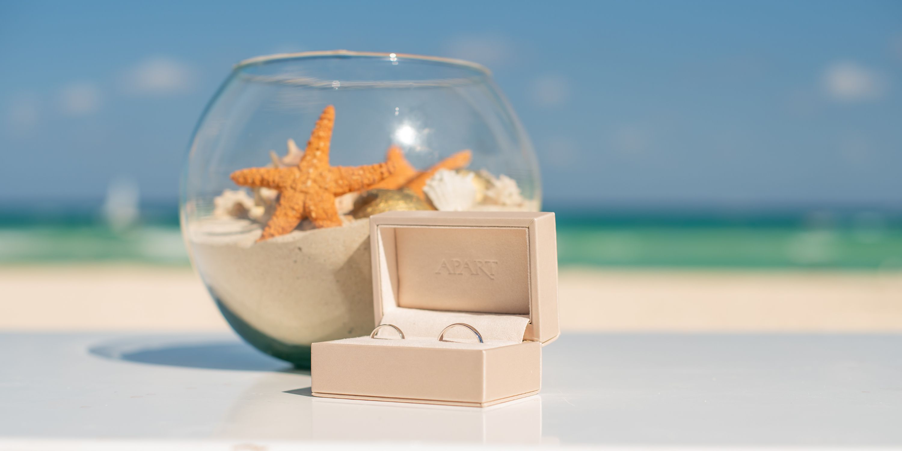 Wedding rings in a box are showcased in front of a bowl with sand and seashells at a destination wedding.