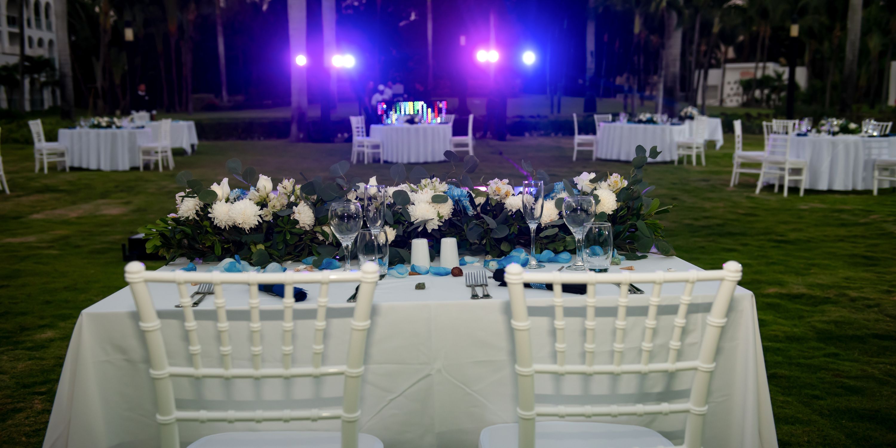 White chairs and a flower-adorned table set for a destination wedding at Hotel Riu Palace Mexico outdoors.