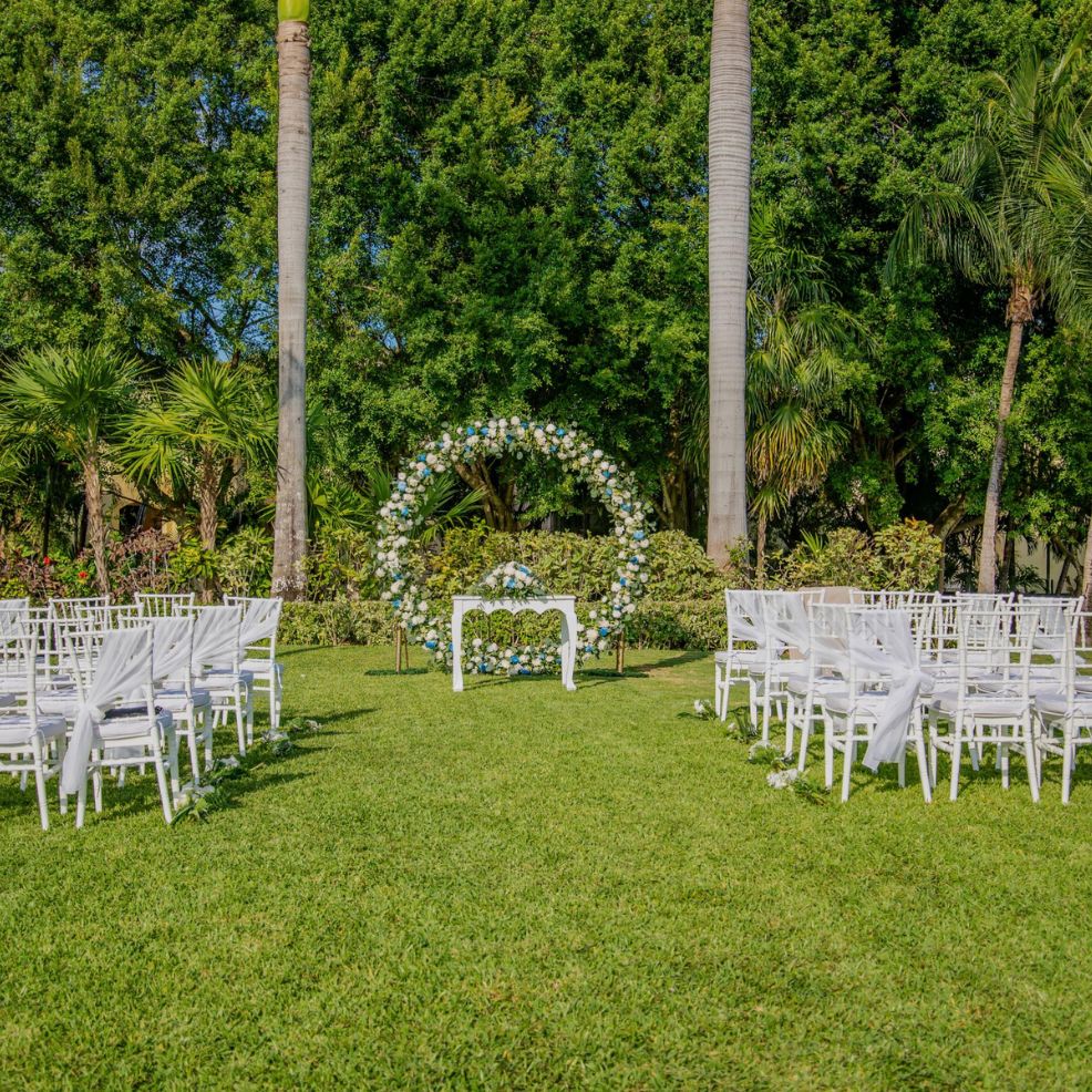 Destination wedding ceremony featuring white chairs, a floral arch, and a small table on grassy lawn.