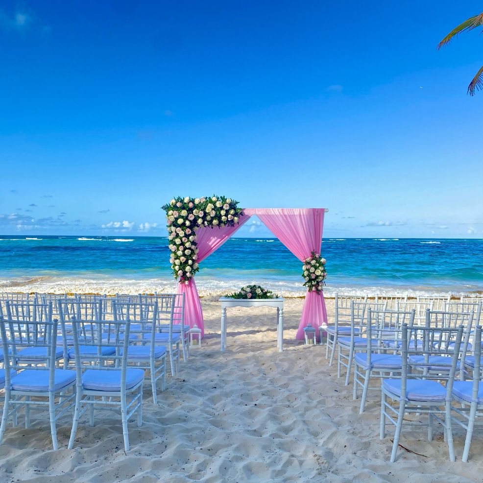 White chairs and a pink floral arch arranged for a destination wedding on a sandy beach by the ocean.