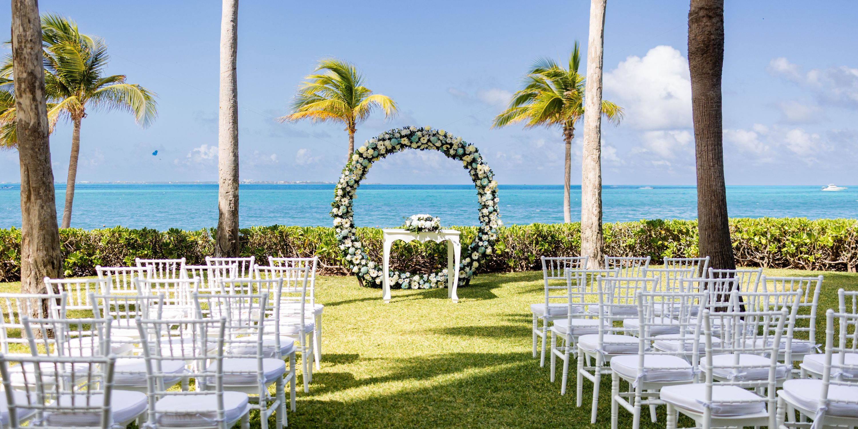 Destination wedding setup at Hotel Riu Palace Peninsula featuring white chairs and a floral arch by the ocean.