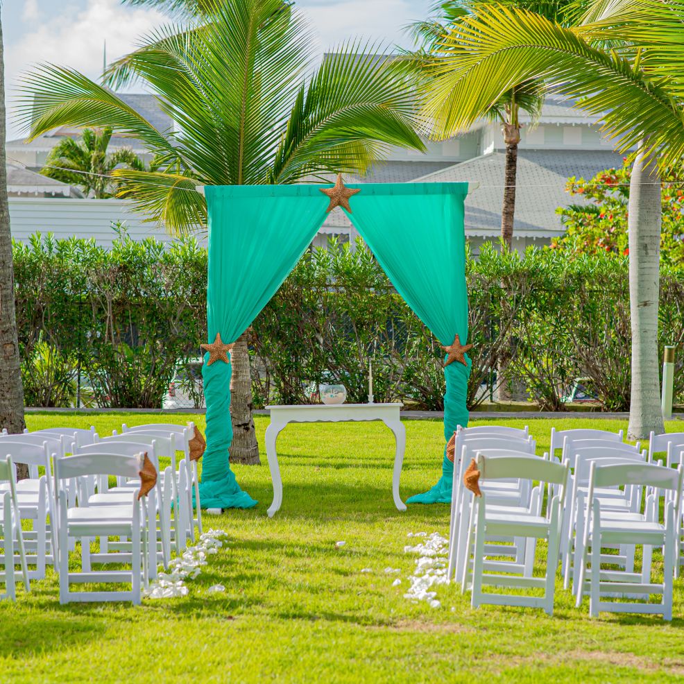 Destination wedding ceremony arrangement featuring rows of white chairs and a turquoise-draped arch.