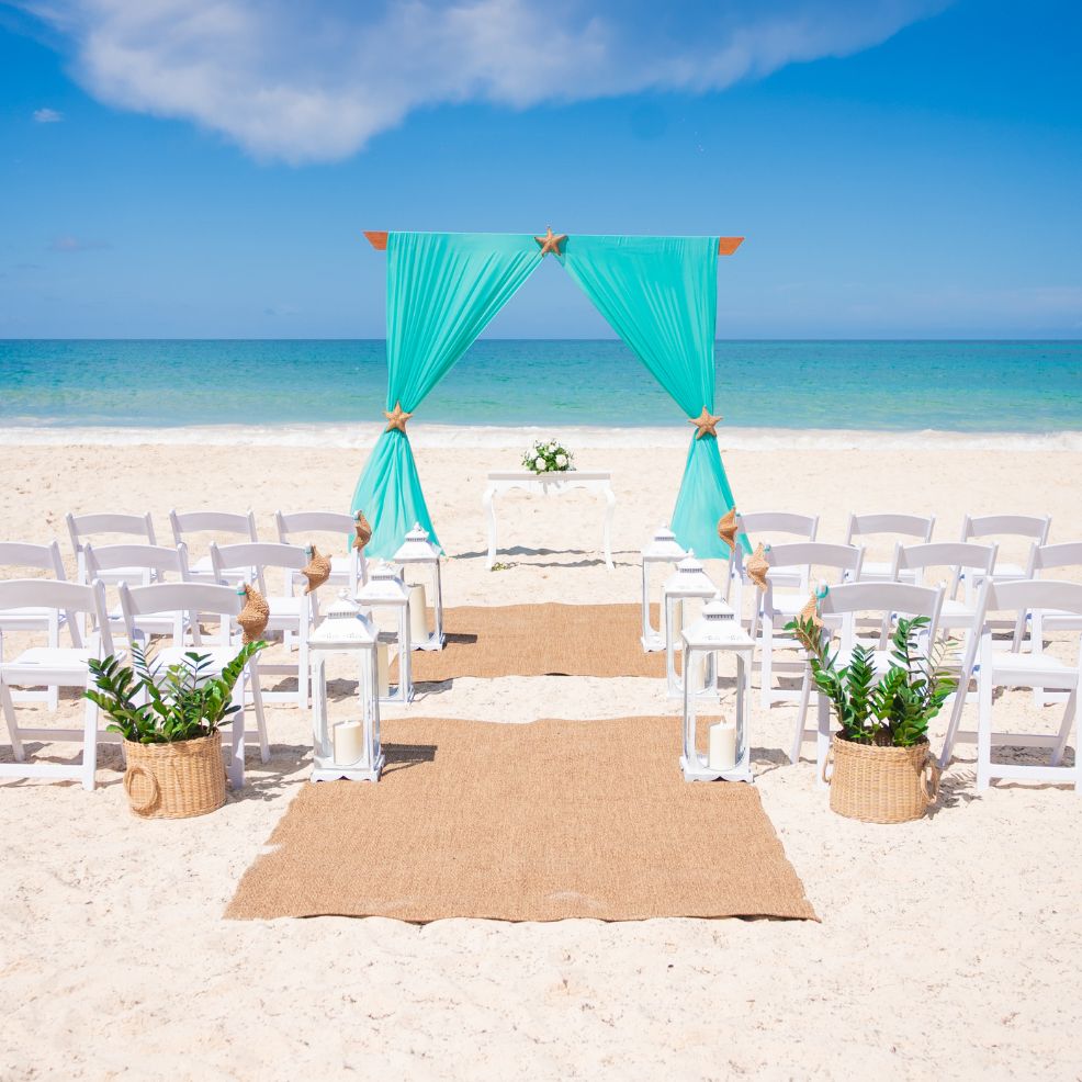 Destination wedding setup on the beach with white chairs, turquoise drapes, and a small floral altar.