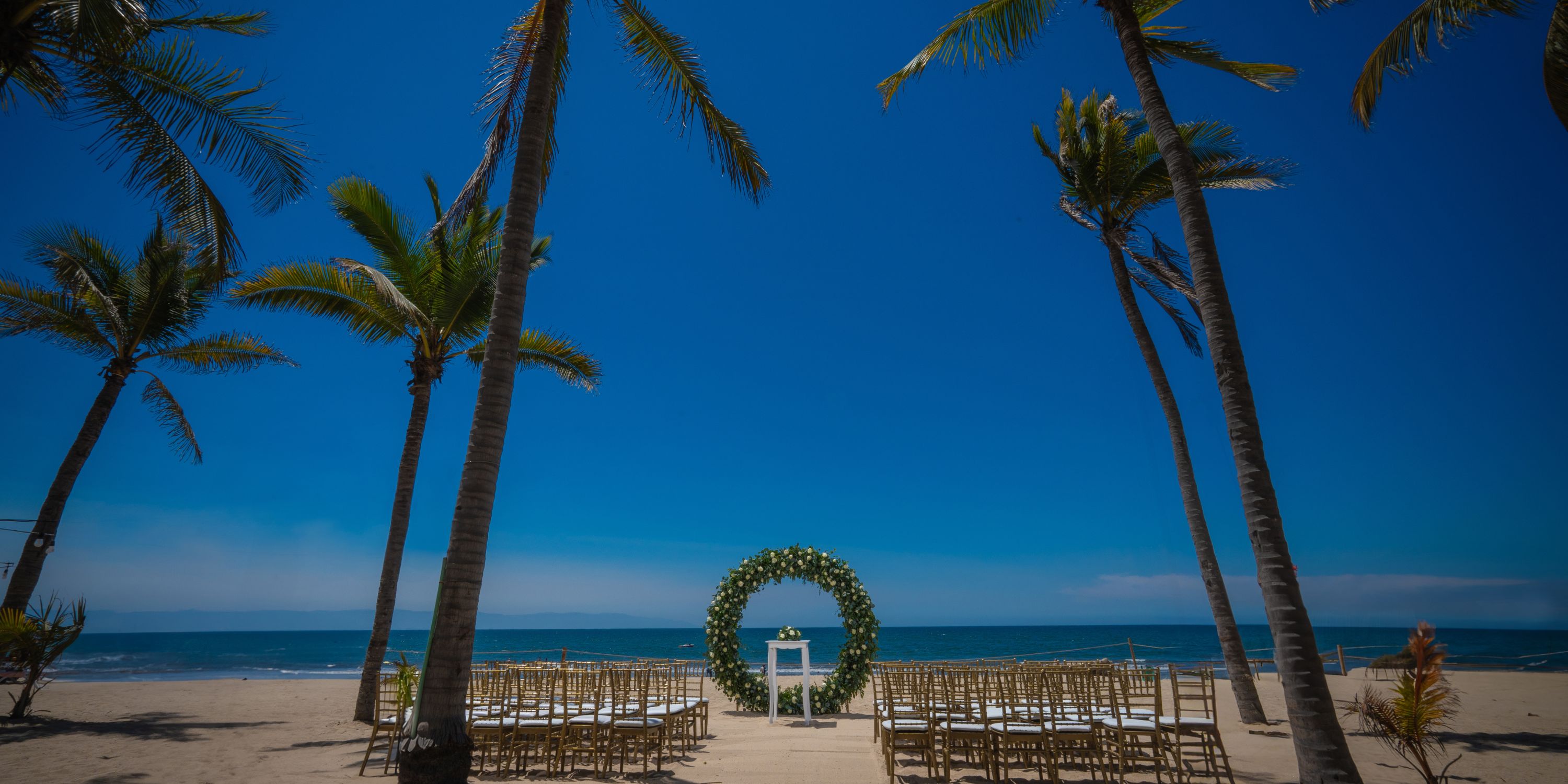 Chairs set up for a destination wedding face a floral arch on the sandy beach at Hotel Riu Vallarta.