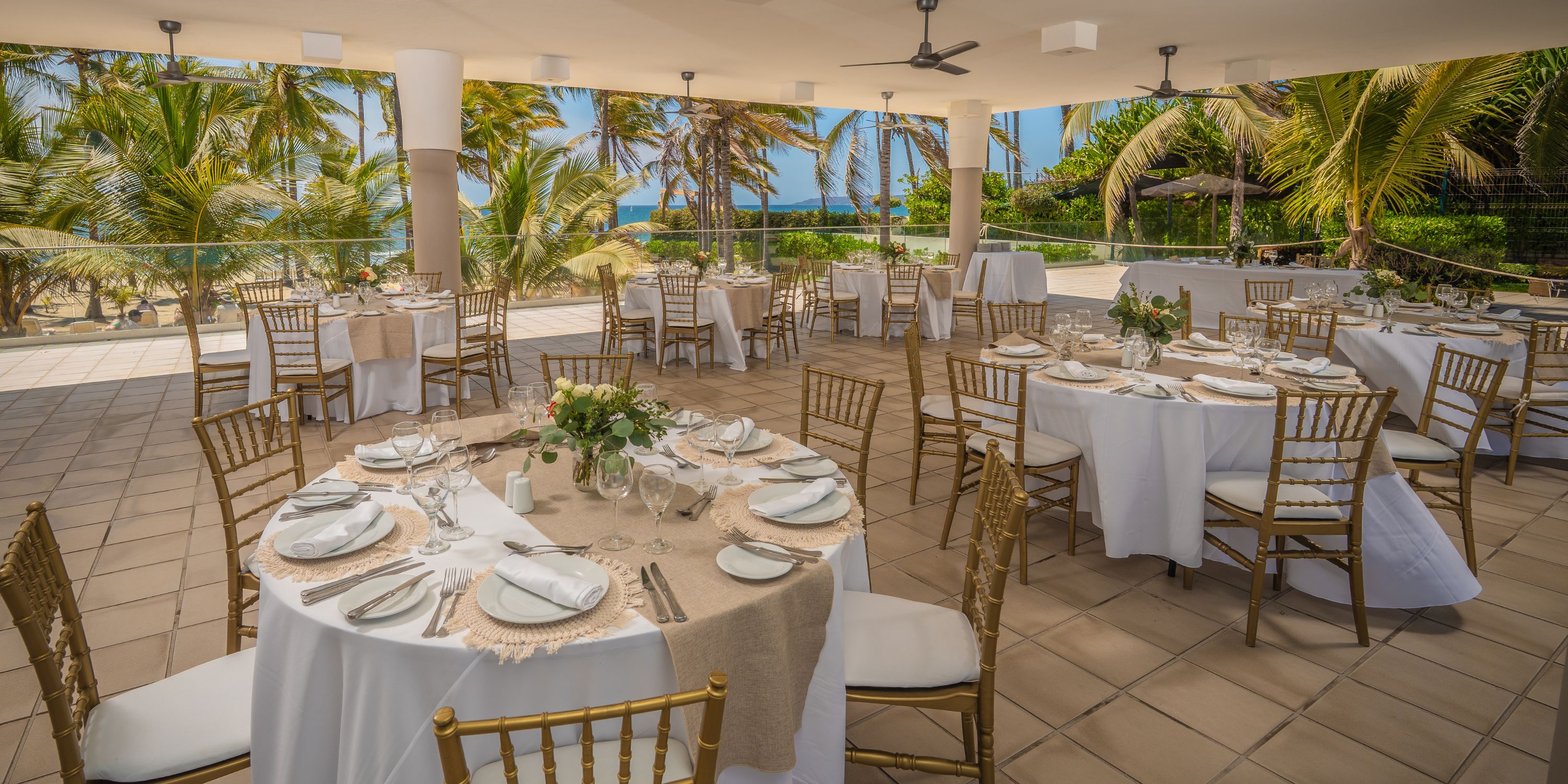 Hotel Riu Vallarta&rsquo;s outdoor banquet area is set with round tables for a destination wedding by palm trees.