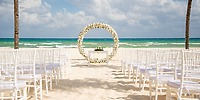 White chairs are arranged for a destination wedding by a floral arch on the beach at Hotel Riu Yucatan.