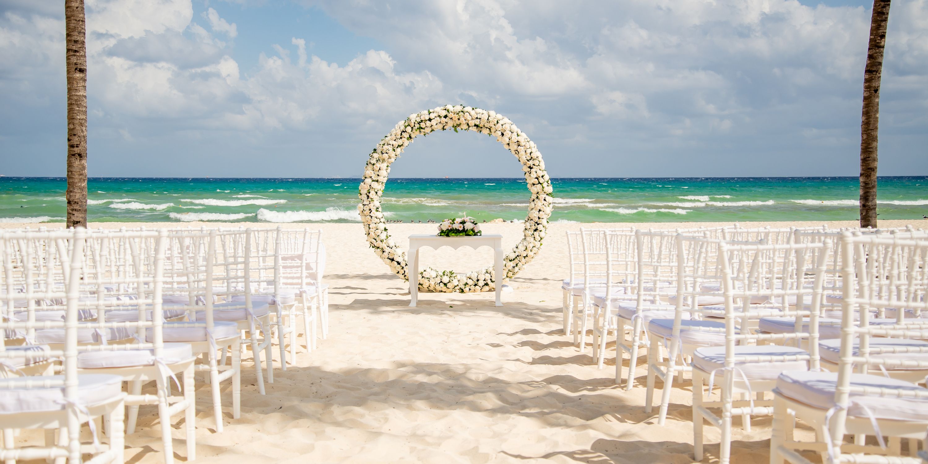 White chairs are arranged for a destination wedding by a floral arch on the beach at Hotel Riu Yucatan.