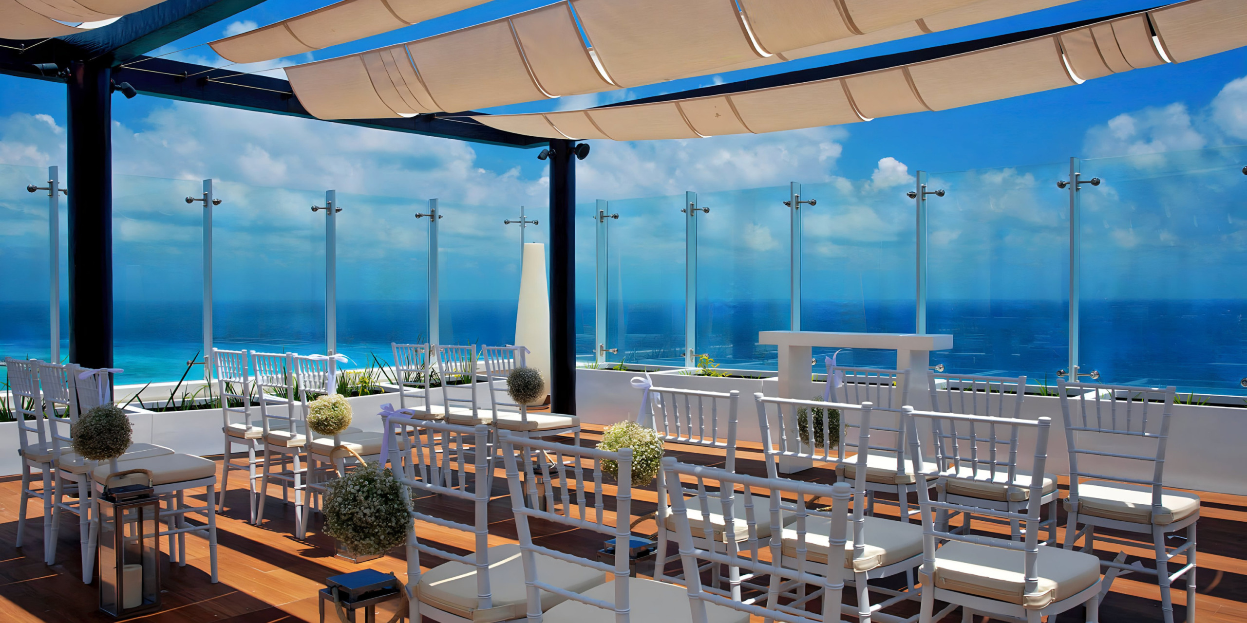 Outdoor seaside destination weddings dining area with white chairs and tables under a canopy, overlooking a clear blue ocean under a bright sky.