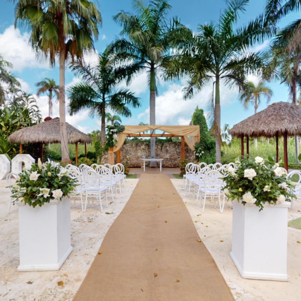 Destination wedding ceremony arranged outdoors with rows of white chairs, floral decor, and palm trees.