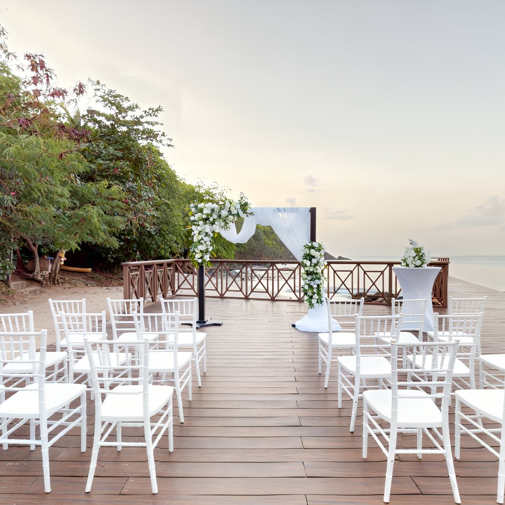 Destination wedding ceremony on a wooden deck by the beach, featuring white chairs and a floral arch.