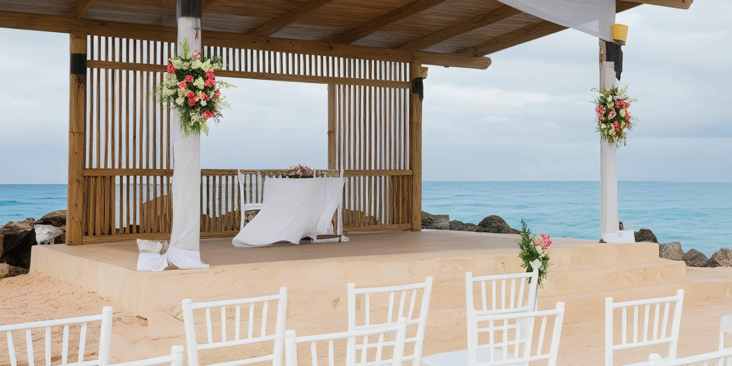A picturesque beachfront wedding scene at Hideaway at Royalton Blue Waters features a charming wooden gazebo adorned with flowers, perfectly positioned to overlook the ocean. Rows of elegant white chairs face the gazebo, ready to accommodate your guests.