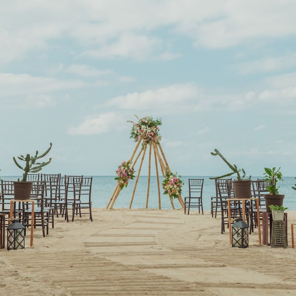 Rows of chairs are set up for a destination wedding, facing a floral arch on the beach by the ocean.