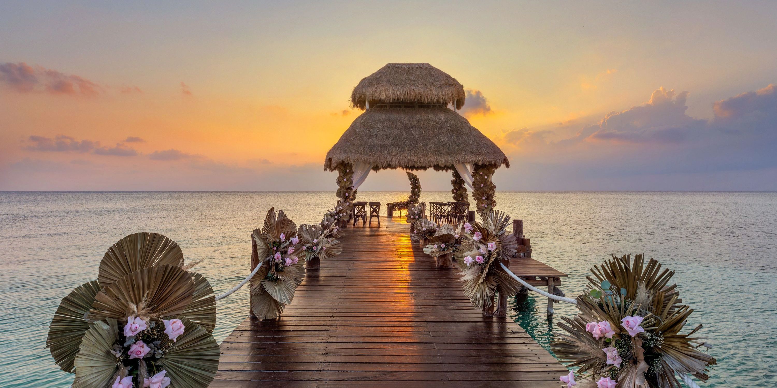 Wooden pier with flowers leads to a thatched gazebo over water, perfect for a destination wedding.