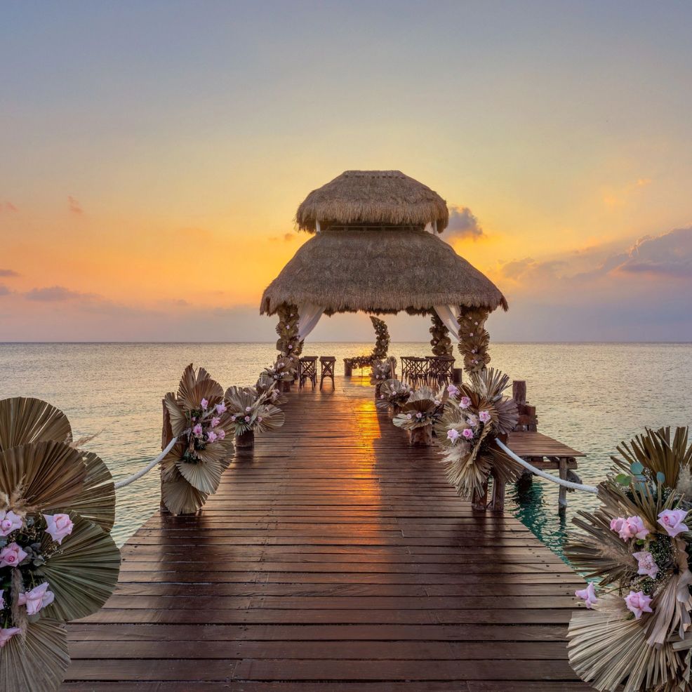 A wooden pier with a thatched-roof hut, perfect for a destination wedding, stretches over the sunset-lit ocean.
