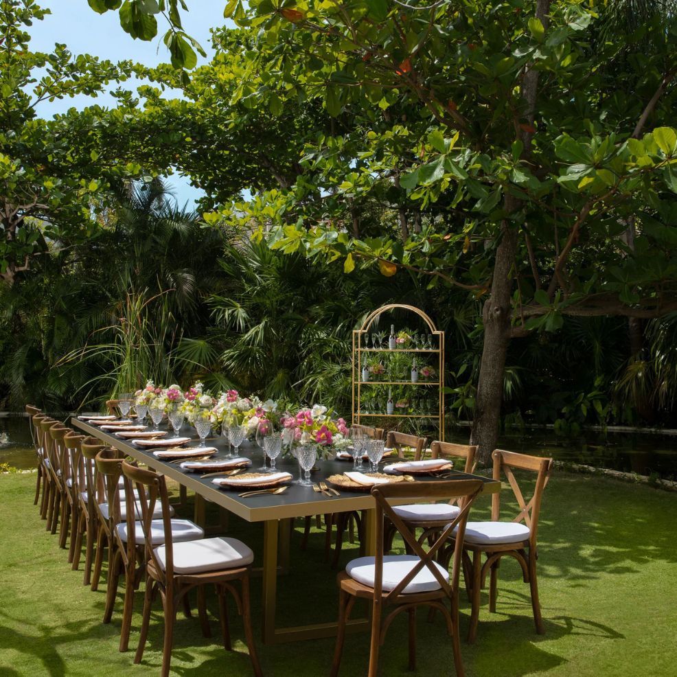 Elegant outdoor dining table with floral centerpieces and wooden chairs set for a destination wedding on grass.