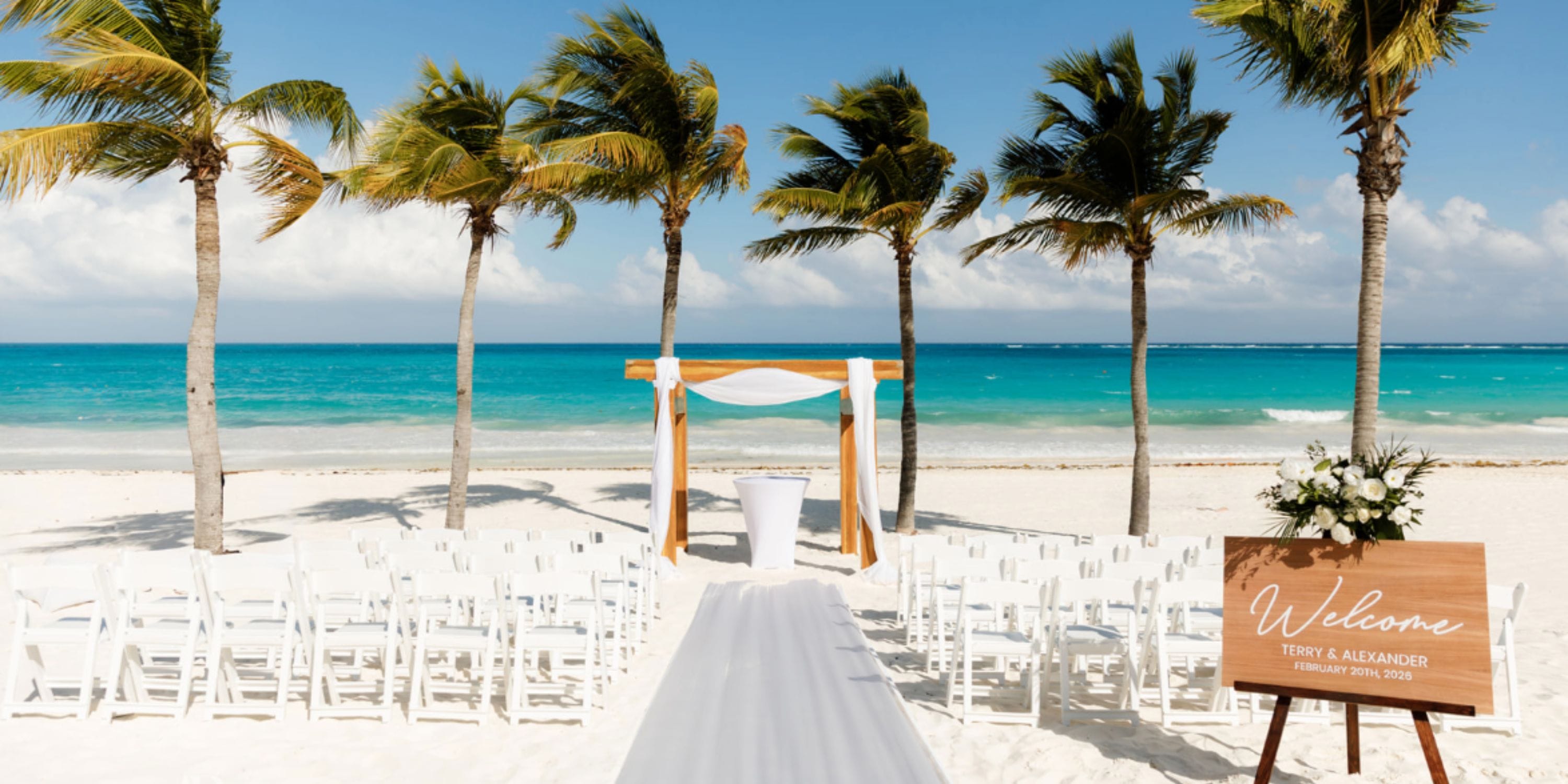 White chairs at a destination wedding arch on sandy Secrets Maroma Beach with palms and ocean views.