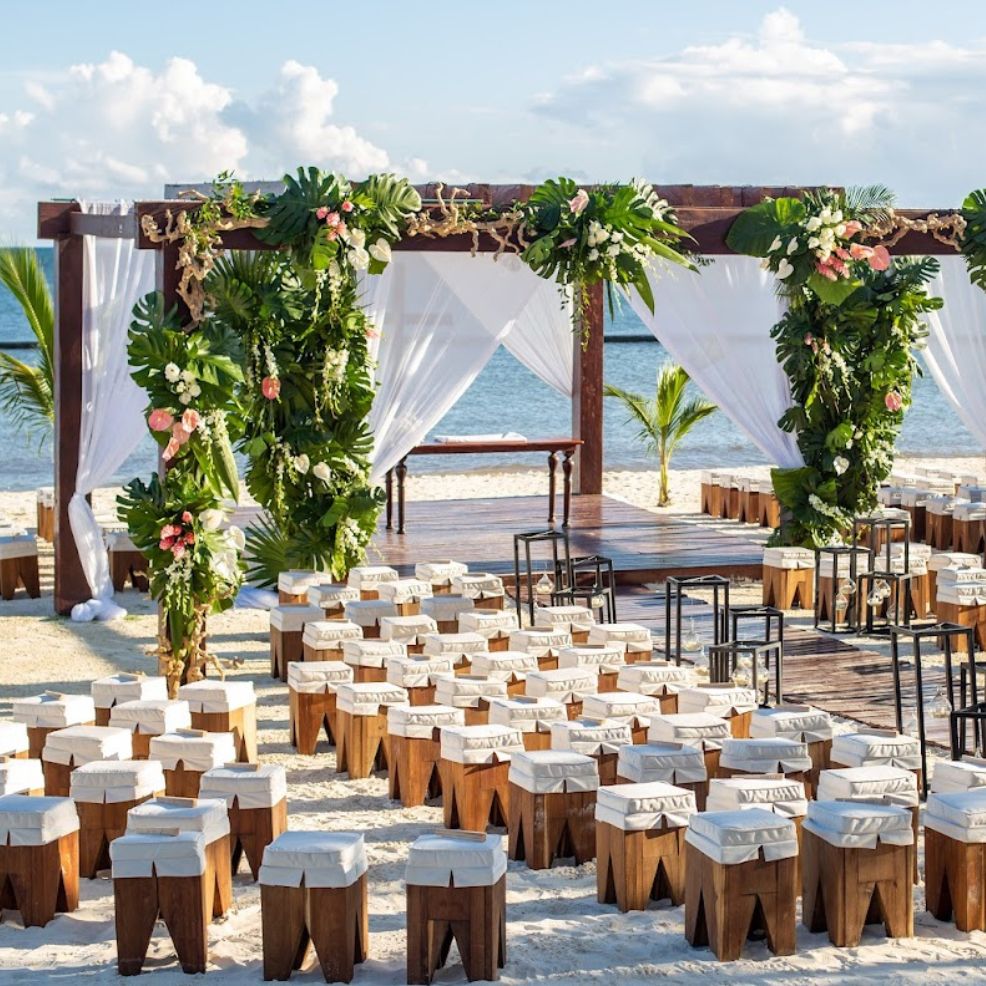 Destination wedding setup on the beach with wooden seats, floral decor, and a canopy overlooking the ocean.