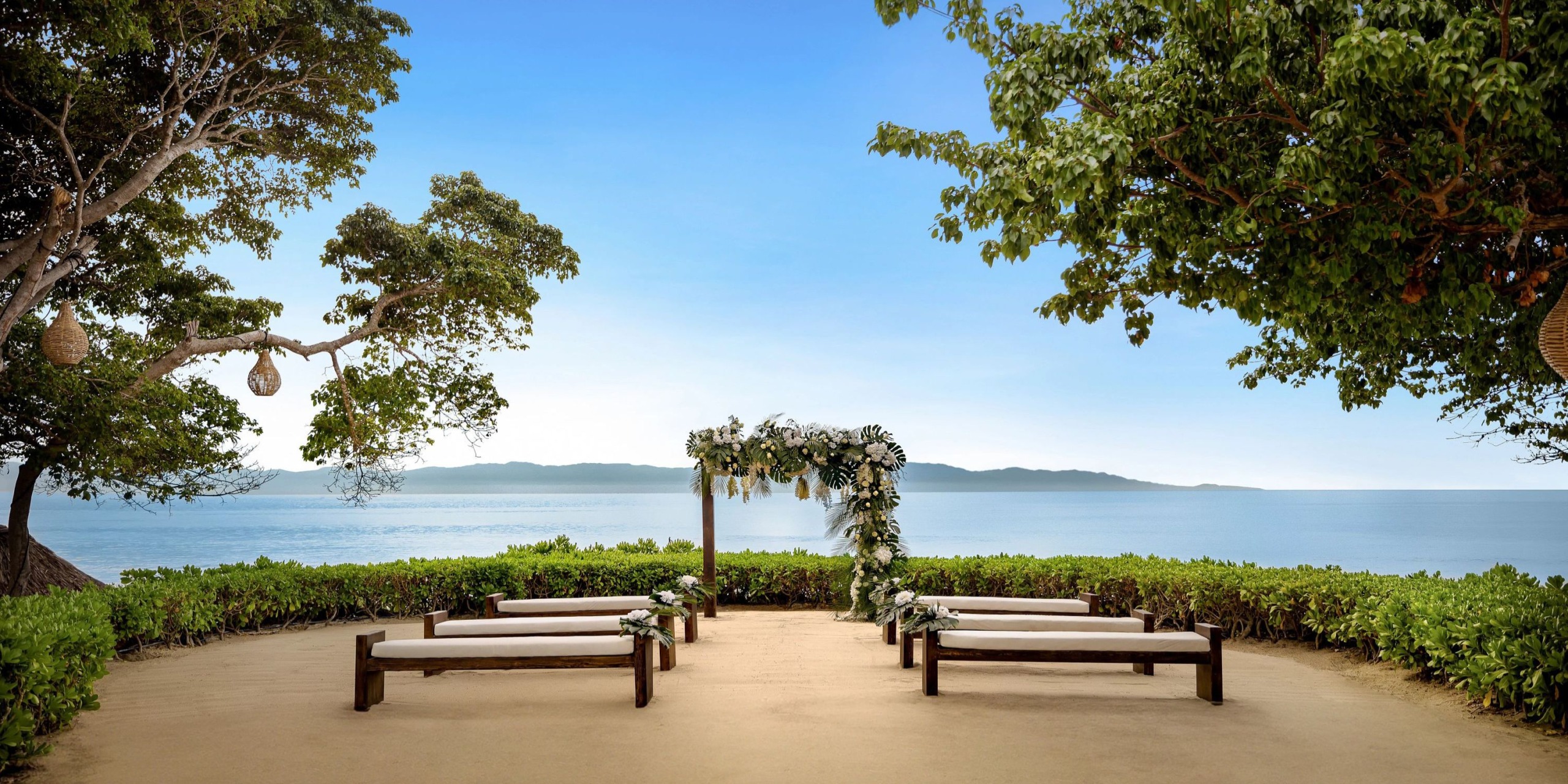 Secrets Bahia Mita offers a picturesque destination wedding setting with a seaside view, featuring an elegant arch and rustic wooden benches nestled beneath a charming tree canopy.